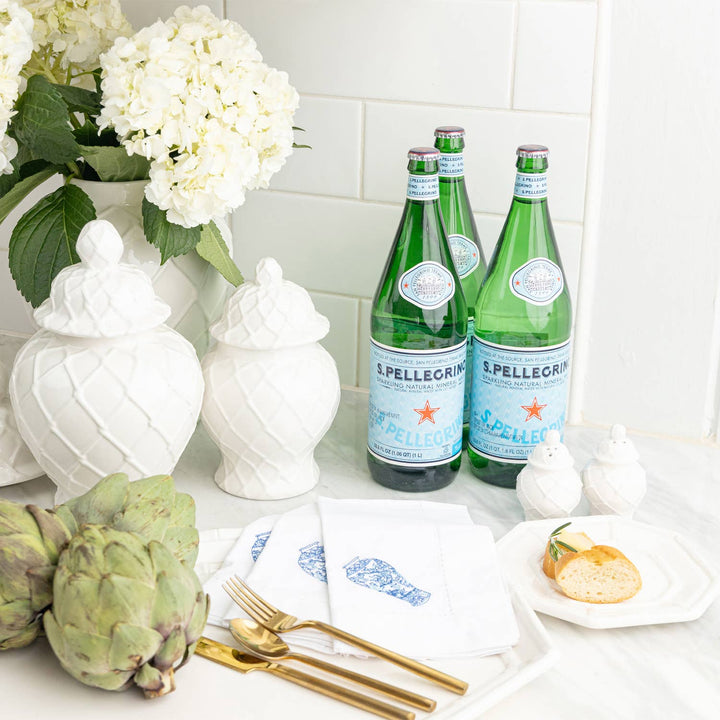 Two bottles of S.Pellegrino on a table with white salt and pepper shakers, white flowers, artichokes, and gold cutlery.