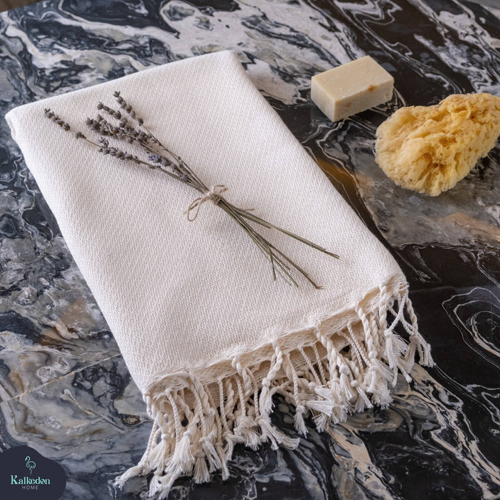 White towel with fringes on a marble surface with lavender, soap, and sponge.