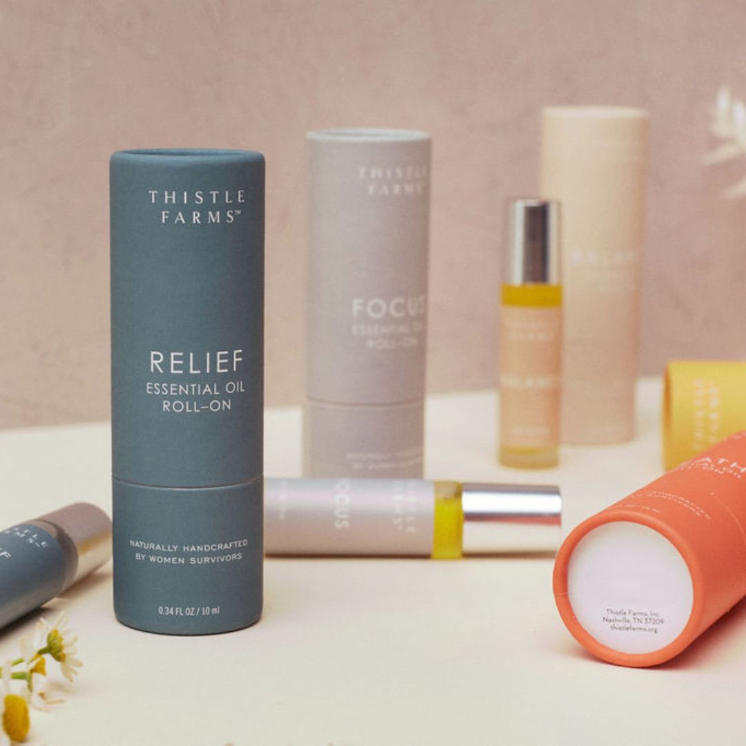 Collection of Thistle Farms essential oil roll-ons on a light surface with a soft pink background.