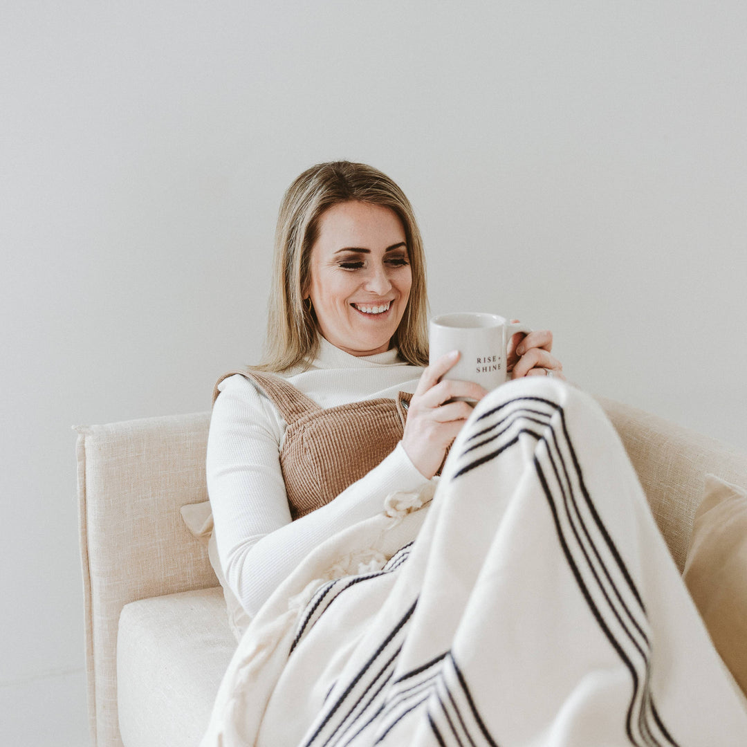 Woman sitting on a couch holding a mug and wrapped in a blanket, smiling.