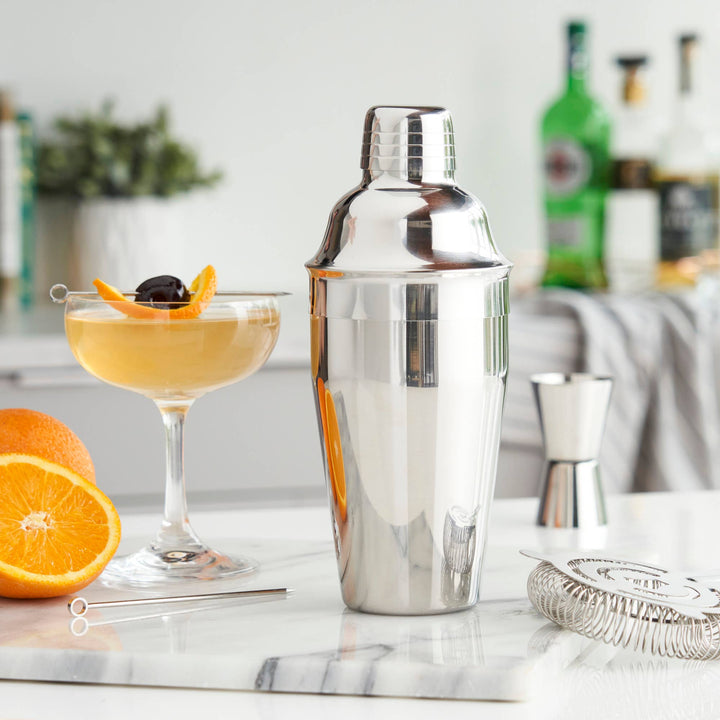 Cocktail shaker with a cocktail glass containing an orange and cherry garnish on a marble surface.