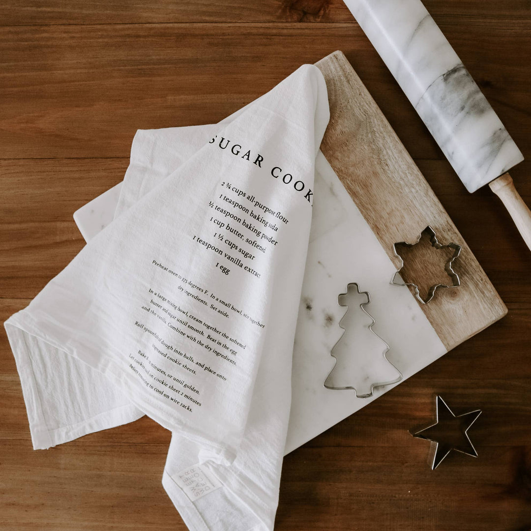 White kitchen towel with text, wooden cutting board with cookie cutters, and rolling pin on a wooden surface