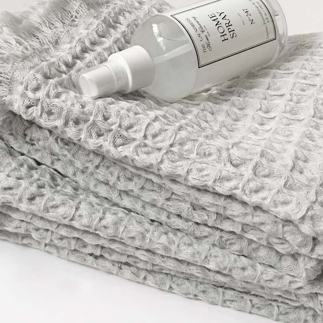Grey textured hand towel with fringe sitting on a white surface and a home spray bottle on top. 