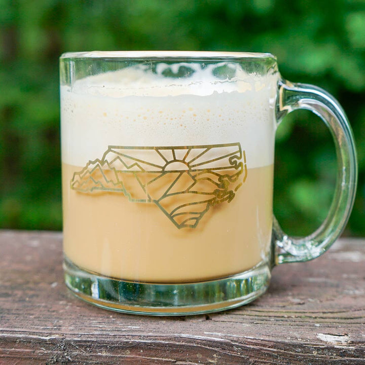 Glass mug with a geometric design in the outline of North Carolina on a wooden surface with a green blurred background.