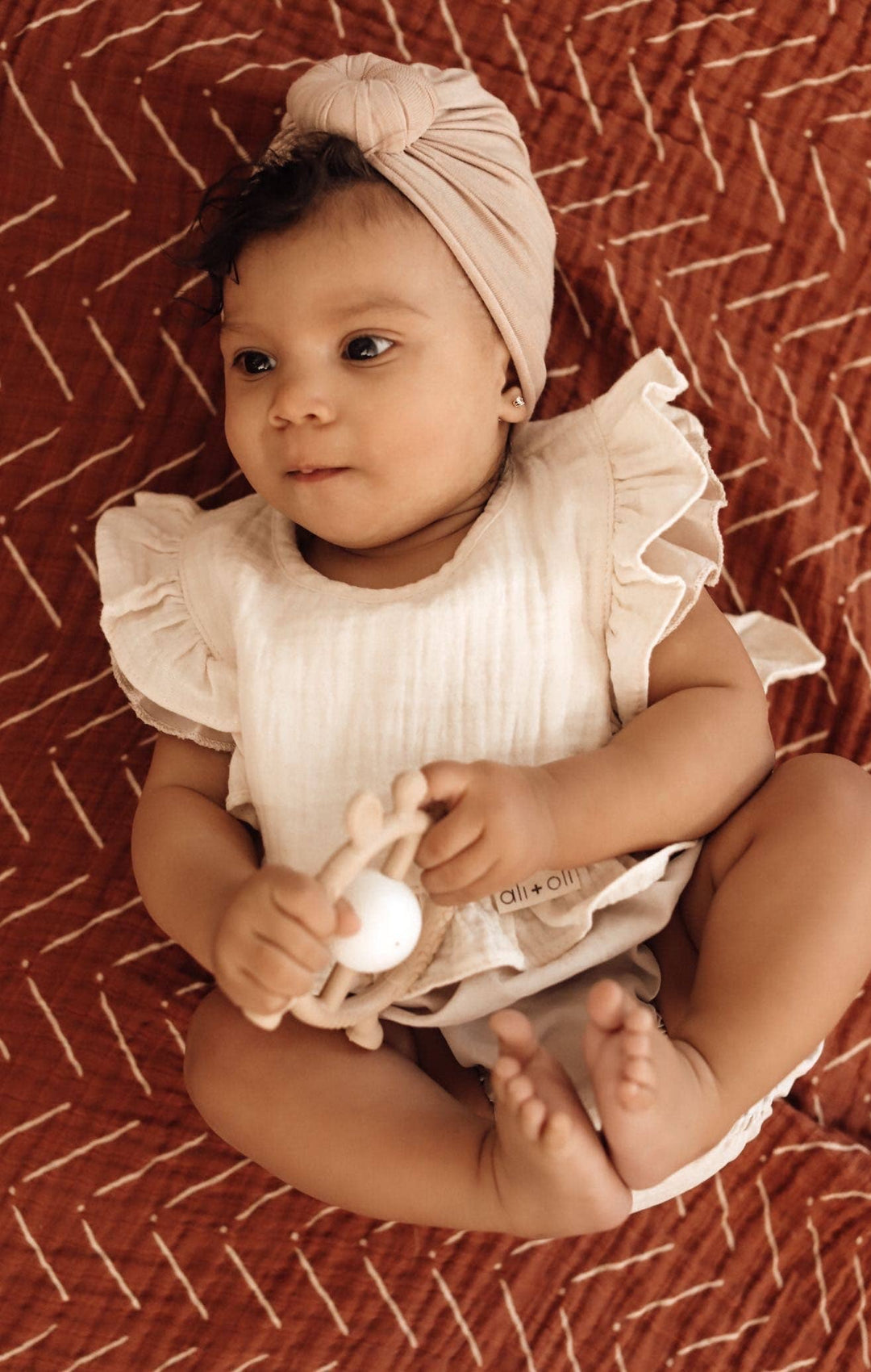 Baby wearing a white ruffled outfit and headband, sitting on a patterned surface.