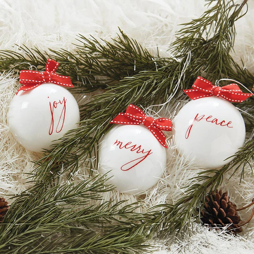 Three white Christmas ornaments with red bows and festive words on a bed of greenery.