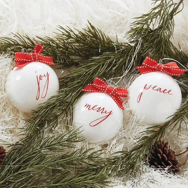 Three white Christmas ornaments with red bows and festive words on a bed of greenery.