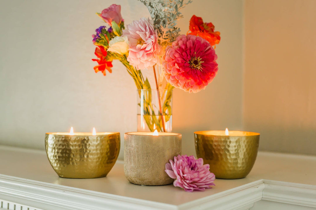 3 different gold textured candles on a white surface with a colorful flower vase behind. 