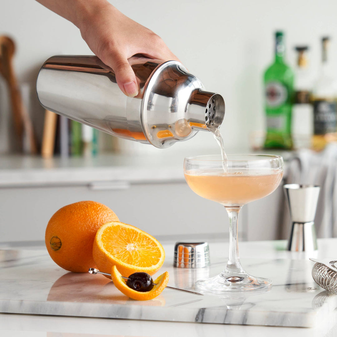 Person pouring a cocktail from a shaker into a glass with oranges and a bar setting in the background.