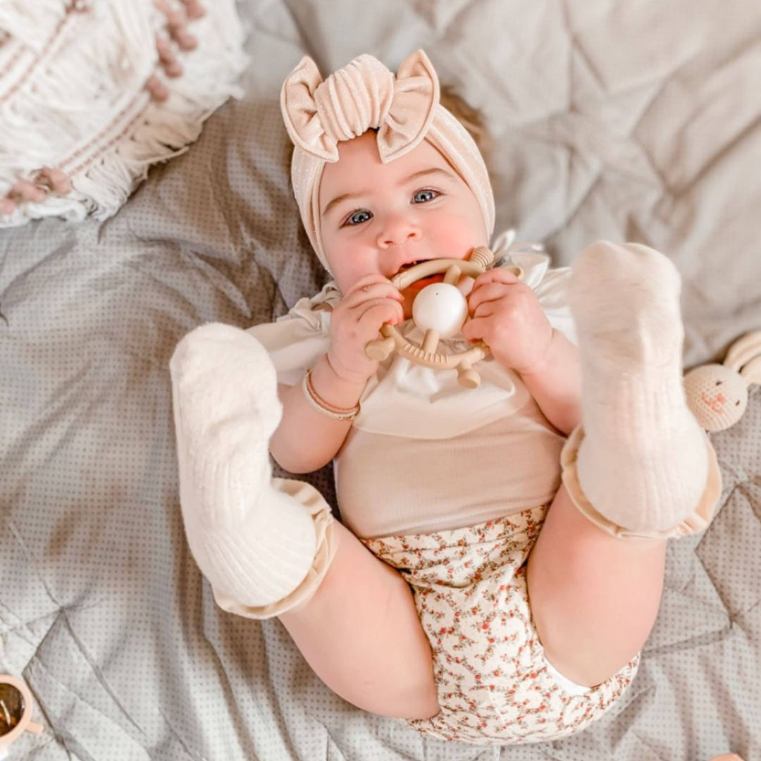 Baby lying on a textured surface wearing a floral outfit and headband, holding a teething ring.