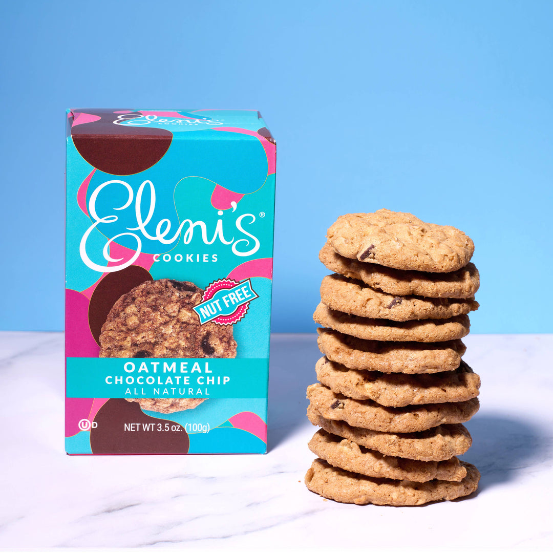Stack of oatmeal chocolate chip cookies next to a box labeled 'Eleni's Cookies' on a white surface with a blue background.