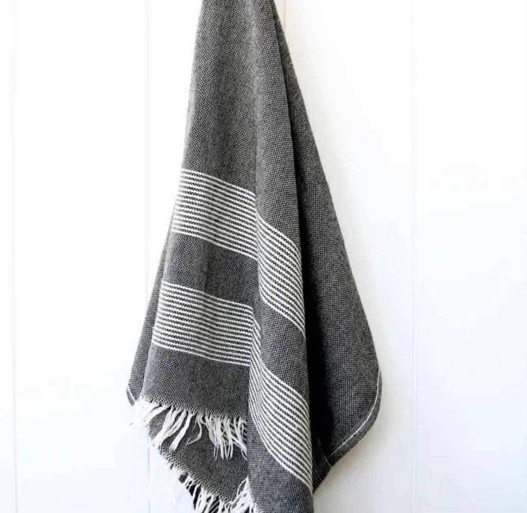 Dark grey towel with white stripes and fringe hanging upon a white surface. 