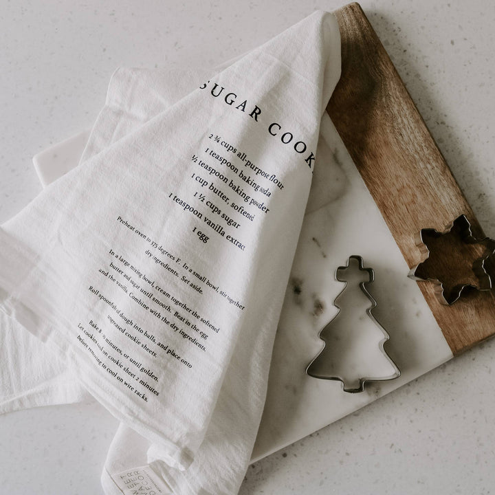 White kitchen towel featuring sugar cookie recipe on a wooden cutting board, and metal cookie cutters on a light surface.