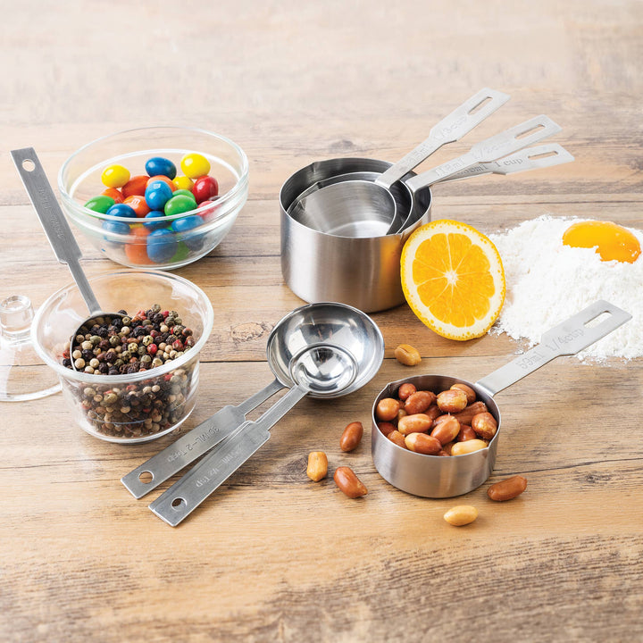 Various measuring cups and spoons on a wooden surface with bowls of candies, nuts and spices. 
