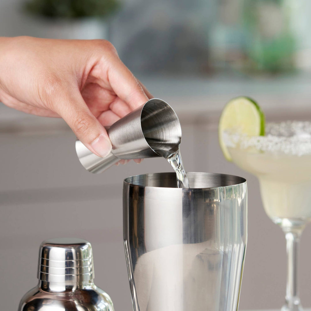 Hand pouring liquid from a metal jigger into a cocktail shaker with a blurred bar setting in the background.