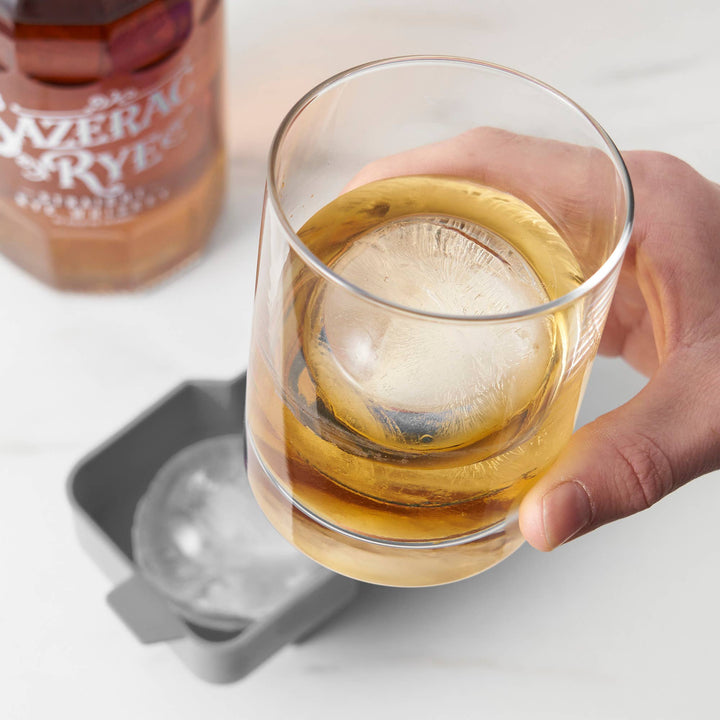 Hand holding a glass of whiskey with a large ice cube, next to a bottle of Sazerac Rye on a light surface.