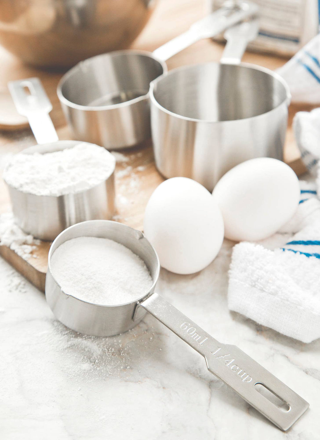 Different sizes of measuring cups filled with flour with eggs, towels and a bowl in the background. 