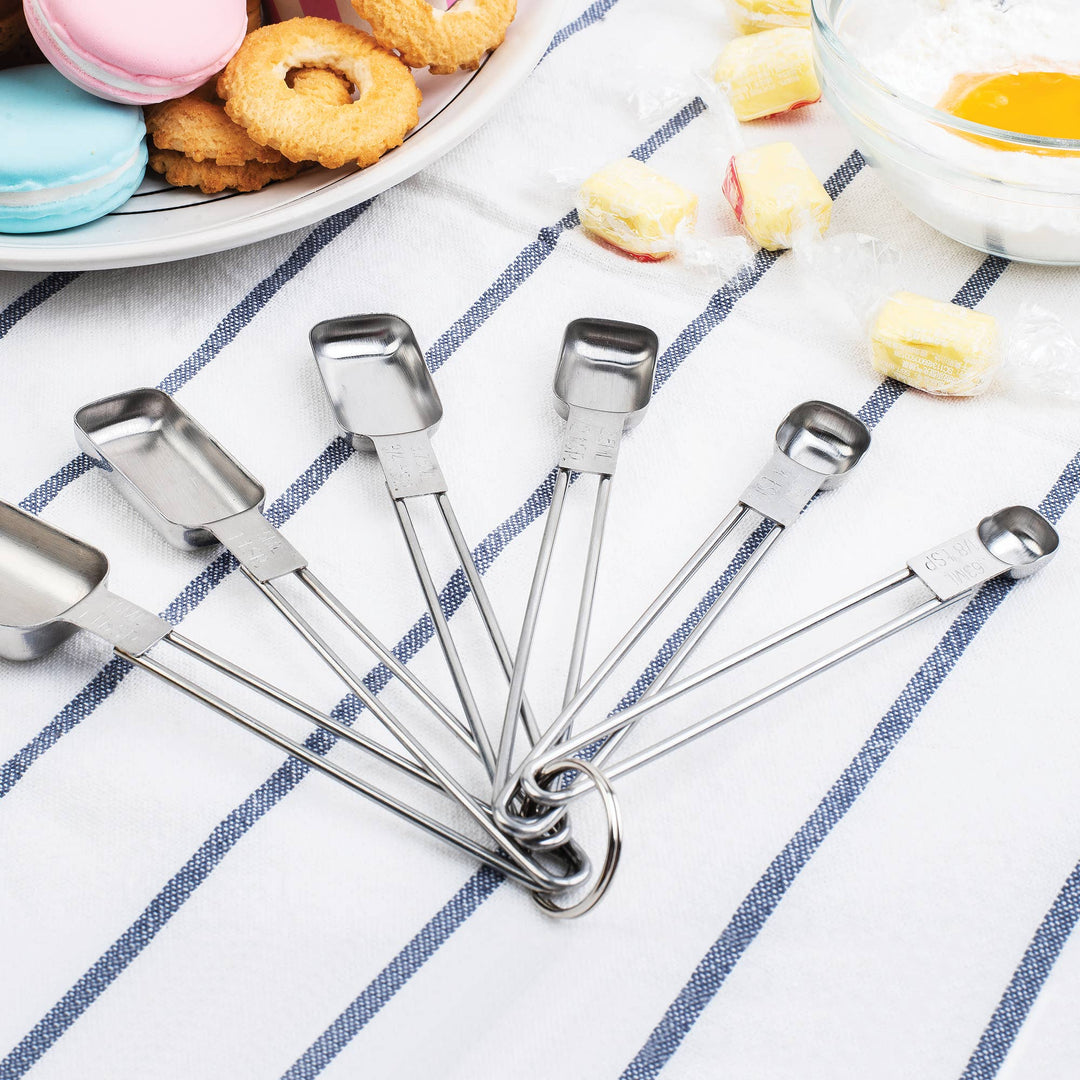 Various sizes of stainless steel measuring spoons on a striped towel surface with snacks and sweets in the background. 